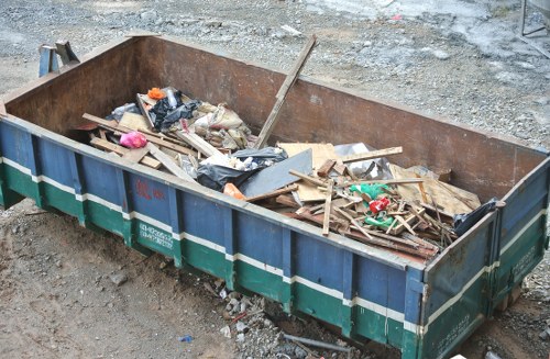 A large, rectangular metal skip is positioned on a gravel and dirt surface, filled with mixed construction and household waste including wooden planks, plastic bags, cardboard, and various small debris. The skip's exterior is painted in green with a blue stripe and has visible signs of rust and wear, indicating frequent use for rubbish collection. It is situated outdoors, likely on a commercial or construction site, with a rough, uneven ground surrounding it. The environment suggests an active waste disposal area, possibly corresponding with rubbish removal services by Commercial Waste Clapham, catering to business waste collection needs in the local area. Natural daylight illuminates the scene, highlighting the textures of the various materials inside the skip, with some cleanup debris scattered nearby, reinforcing the context of waste management in a contemporary urban setting.