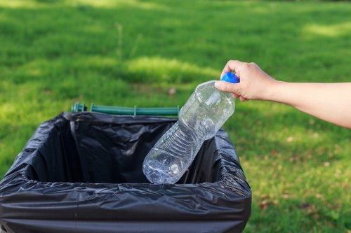 A person’s hand is seen holding a clear plastic water bottle with a blue cap, about to place it into a black plastic rubbish bin. The bin is situated outdoors on a paved surface or driveway, with a background of well-maintained green grass and trees, suggesting a garden or park area. The water bottle appears to be partially crumpled and is made of transparent PET plastic, reflecting natural daylight. The scene indicates waste collection or rubbish disposal activity, which is relevant to professional rubbish removal services provided by Commercial Waste Clapham, serving the Clapham area. The environment is bright, with the focus on the act of recyclable waste being properly disposed of, emphasizing the importance of responsible rubbish management in a suburban or urban setting. The image captures the clear process of environmental responsibility within a residential or commercial outdoor space, aligning with the company's role in local waste management solutions.