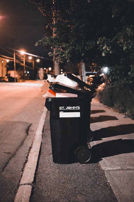 A professional waste collection worker wearing a high-visibility orange vest and blue uniform is placing a blue wheelie bin into the back of a white garbage truck parked on a cobbled urban street. The