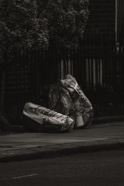 Three large black rubbish bags made of thick plastic and slightly crinkled, are placed on the pavement in front of a black metal fence with vertical bars. The bags appear to contain household waste an