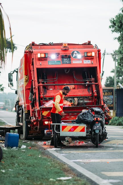 A large pile of uncollected rubbish, composed mostly of black plastic trash bags, is situated in front of a red metal door on an exterior wall. The bags are filled with various waste items and are sta
