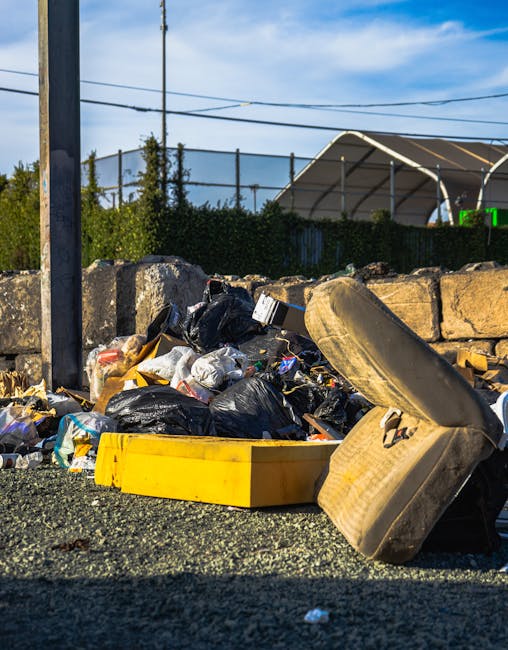 A waste collection worker dressed in a red and yellow uniform is standing at the rear of a large red rubbish truck on a street. The truck's back panel is open, revealing the compaction mechanism and i