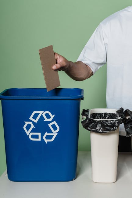 A person dressed in a white shirt is seen holding a rectangular piece of brown cardboard above a blue recycling bin with a white recycling symbol on the front, positioned on a light-colored surface ag